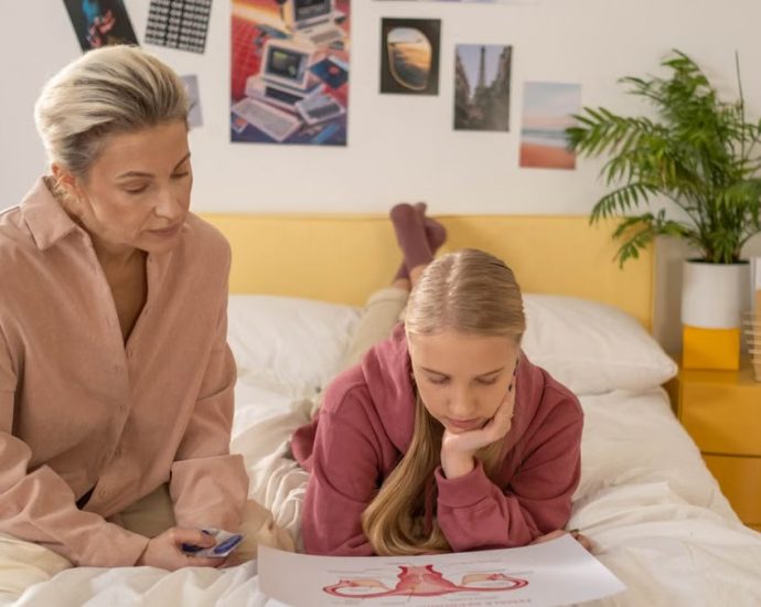 A mother and daughter talk over illustrated materials in a cozy bedroom setting.