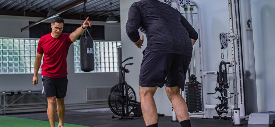 Back view of muscular man in sportswear doing exercises with trainer in modern fitness club